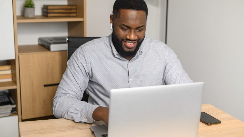 Man sitting as desk, smiling while looking at a laptop