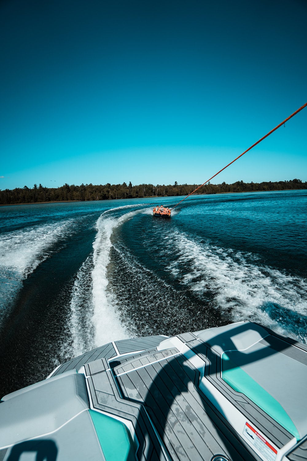 Photograph of the back of a boat, pulling people on a tube, on a lake. Edited, color and contrast are increased with an emphasis on orange and teal.