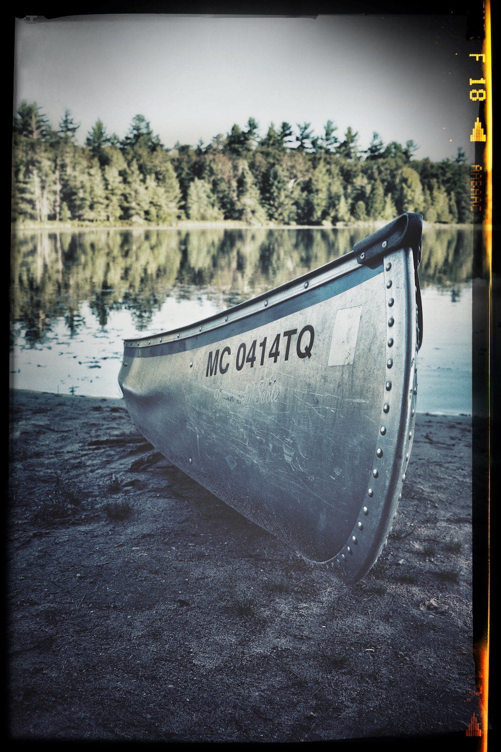Photograph of a aluminum canoe the bank of a lake, edited, color and contrast are increased. Has a film boarder