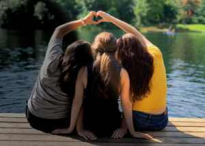 Three girls sitting on a dock making a heart shape with their hands. Half of the image is dark and black and white, the other half is in color.