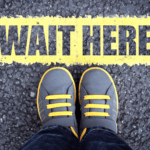 a line on a sidewalk with the words 'wait here' and someone standing behind the line.